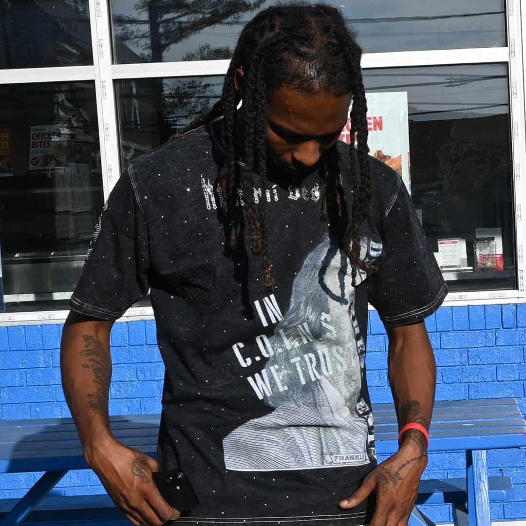 Person wearing a black t-shirt with text, sitting on a blue bench in front of a store.